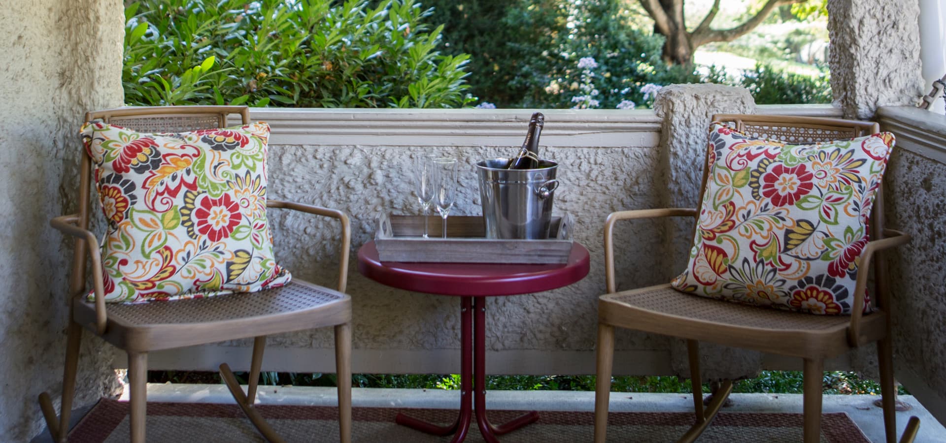 A cozy patio scene featuring two chairs with colorful cushions and a small table holding a tray, wine glasses, and a champagne bucket.