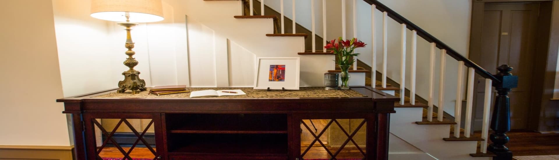 A wooden table with a lamp, flowers, and framed art sits beside a staircase in a well-lit entrance area.