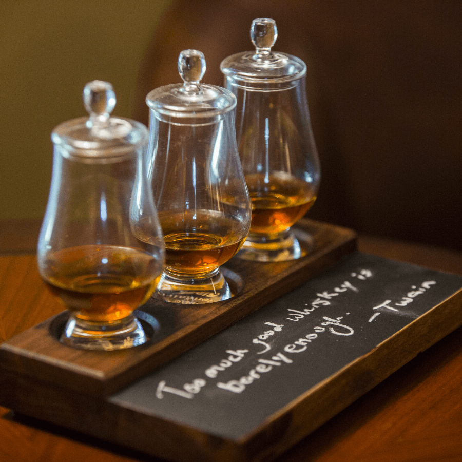 A wooden flight board with three whiskey glasses and a quote by Twain on a chalkboard.
