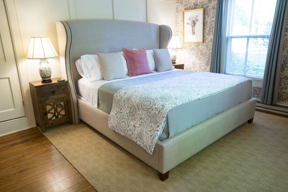 A cozy bedroom featuring a queen-sized bed with decorative pillows, a patterned blanket, a bedside lamp, and floral wallpaper.