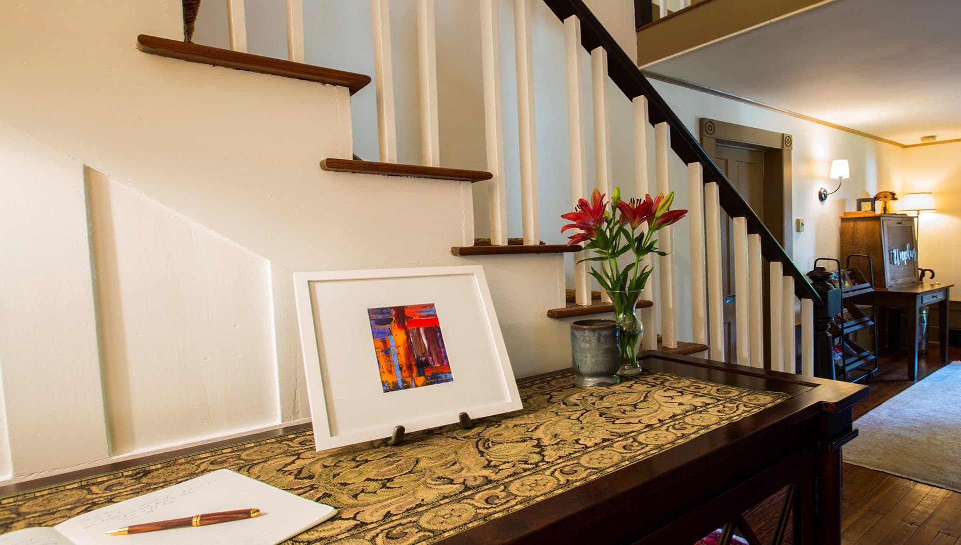 A cozy entryway table decorated with a framed artwork, fresh flowers, and a notepad.